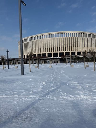 The image depicts a winter scene outside the FC Krasnodar Stadium in Plodorodny, Russia. The stadium, a large, circular structure with a textured, light-colored facade of vertical ribbed panels and numerous rectangular windows, dominates the background under a clear blue sky. In the foreground, a wide expanse of ground is heavily covered in fresh, undisturbed snow, with clear human footprints forming paths and faint tire tracks visible closer to the camera. Several tall, slender, dark grey lampposts with modern LED fixtures are positioned across the scene, one prominently in the immediate foreground, casting long shadows. Numerous small, deciduous trees with bare branches are sparsely planted throughout the snow-covered area, each supported by a light-colored, cuboid base or planter. Dark-colored pergolas or covered walkways are visible in the midground, leading towards the stadium entrance areas. No individuals are visible in the frame. The scene is illuminated by bright daylight, indicating clear weather.