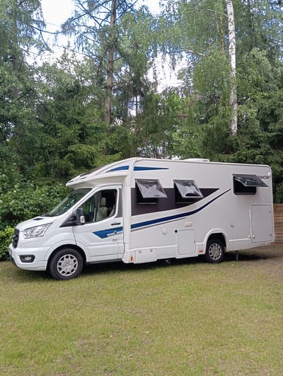 A white recreational vehicle (RV) is parked on a grassy area with a dense forest in the background. The RV has tinted windows with the top part of some of them open, and blue and dark gray accents on its exterior. The front of the RV is a white Ford Transit van. The scene is illuminated by daylight, suggesting it is daytime. The location is identified as Mołowiste, Poland. There are no people visible in the image. The RV appears to be stationary.