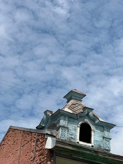 A close-up, low-angle shot captures the top of a weathered brick building and a distinctive blue-painted wooden mansard roof structure against a cloudy sky. The brickwork, a warm reddish-pink hue, exhibits signs of age and some damage. The mansard roof is ornate, featuring peeling blue paint, arched window openings, decorative carved elements, and a tiered cap. The weathered roof shingles are visible on the mansard. Several thin electrical wires stretch horizontally across the frame, partially obscuring the sky and the upper parts of the building. The sky is filled with soft, white cumulus clouds against a light blue backdrop. The scene appears to be in the city of Nogornyi, Russia. There are no people or active events depicted in the image.