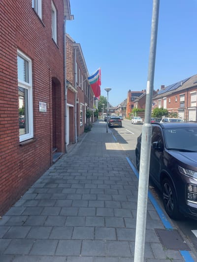 This is a sideways, eye-level shot of a residential street in Venlo, Netherlands, on a clear, sunny day. The scene is dominated by red brick buildings with white-trimmed windows and solar panels visible on some rooftops. A grey metal pole runs vertically through the left side of the frame, obscuring part of the sky.
In the foreground, a section of a red brick building is visible, with a white window reflecting the sky. Further down the street, a flag with red, white, green, and blue stripes is flying. Cars are parked along the right side of the street, with blue markings on the curb indicating parking spots. The pavement is made of grey rectangular tiles.
The time of day appears to be midday or early afternoon, given the bright sunlight and the long shadows cast by the buildings. The weather is clear and pleasant. A small, white street sign is affixed to the brick building, partially obscured by the pole. The overall atmosphere is one of a quiet, everyday street scene in a European town.