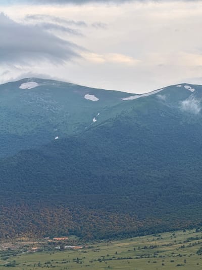 The image depicts a vast, mountainous landscape. The upper portion of the image is dominated by a soft, cloudy sky with subtle gradients of gray and pale yellow. Below the sky, a large, green mountain range stretches across the frame. Patches of snow are visible on the higher elevations of the mountain. The middle section of the mountain is densely covered in dark green forest.

The lower part of the image transitions to a wide, green valley. Scattered trees dot the valley floor. In the mid-ground of the valley, there are several structures that appear to be buildings, some with illuminated windows, suggesting human habitation or activity. Power lines are visible stretched across the landscape in the lower portion of the image. The overall impression is one of a natural, rural setting with some evidence of human presence.