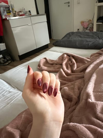 The image primarily features a person's right hand resting on a bed, showcasing manicured nails. The hand has fair skin, and the nails are long, with an almond or oval shape, painted in a deep, glossy reddish-purple or burgundy color that exhibits a metallic or "cat-eye" effect, creating a lighter streak down the center of each nail. The thumbnail appears to have slight wear or a minor chip at its tip.

The hand is positioned over a white fitted bed sheet, which shows some wrinkles. Adjacent to the hand and partially covering the bed is a plush, soft blanket in a pinkish-mauve or light brown hue, appearing thick and textured, likely made of fleece or a similar material, and is somewhat bunched up.

In the background, to the left, is a white nightstand or small cabinet with multiple drawers or doors, featuring thin metallic horizontal and vertical accents that act as handles or dividers. On top of this cabinet, various personal items are visible, including bottles, containers, and a red and white cigarette packet with faint text, possibly "RG," discernible. Further back, a plain white wall is visible, along with what appears to be a light switch or thermostat, and a metallic hook. In the upper left corner, a blurred area suggests shelves or an open cupboard containing items stored in clear plastic bags.

The lighting appears to be natural indoor light, illuminating the scene softly. No specific activity is taking place beyond the hand resting. The overall scene suggests a relaxed, indoor setting, likely a bedroom, in Neunkirchen, Germany.