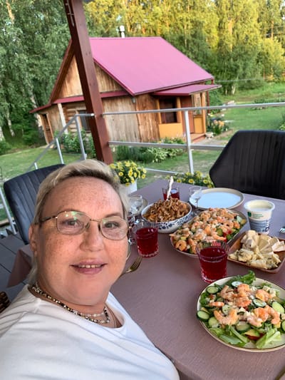 A woman with fair skin and freckles, wearing glasses and a white t-shirt, is taking a selfie outdoors. She is seated at a table laden with food, including a shrimp salad, cheese, and a bowl of what appears to be fried meat or vegetables. There are also glasses of red juice on the table. In the background, a wooden house with a red roof is visible, surrounded by trees and a grassy yard. The scene suggests a meal enjoyed on a porch or deck, with a view of a rural or semi-rural setting in Суроватиха, Russia.