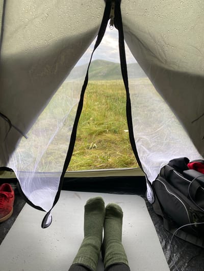 The image is taken from inside a tent, looking out at a landscape. The tent fabric, likely waterproof, shows numerous water droplets on its exterior. The tent opening is partially zipped, with a mesh layer visible. Outside, a grassy, undulating terrain with hills is visible under a cloudy sky.

In the foreground, inside the tent, a person's legs are visible, clad in dark green socks, resting on a light-colored mat. To the left, a red sneaker is partially visible. To the right, a black duffel bag with silver zippers and straps is placed on the tent floor, along with some black cables.

The overall scene suggests a person camping outdoors, possibly in a rural or mountainous area, with the weather being damp. The location is stated to be in the city of Абзаново, Russia, implying this is a view from a campsite in that region.