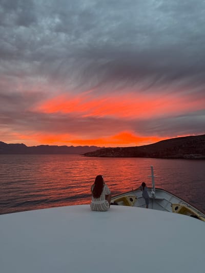 The image depicts a sunset scene from the deck of a boat, possibly a cruise ship, in an unidentified location in Mexico. The sky is filled with grey clouds transitioning into vibrant orange and red hues near the horizon, which illuminates the water below. A dark silhouette of a mountainous coastline is visible along the horizon. Two people are on the deck. One is a woman with long brown hair, leaning against the white railing. The second person is a man standing further back, also near the railing. There are some safety boats on the side.