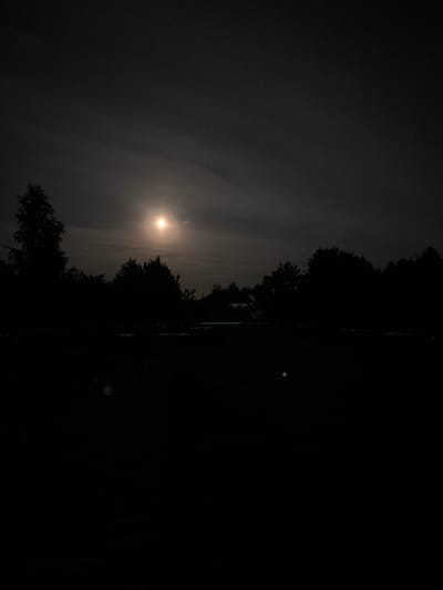 A full moon is visible in the dark sky, partially obscured by clouds, casting an eerie glow over a silhouetted treeline. The foreground is a dark, unlit field, with hints of faint green light suggesting it might be a rural area or park. A few indistinct shapes, possibly structures or vegetation, are visible in the treeline. The overall impression is one of a quiet, moonlit night in a rural setting, possibly in or around Радваніцкі сельскі Савет, Belarus.