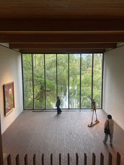 An indoor gallery space at the Louisiana Museum of Modern Art in Humlebæk, Denmark, is viewed from a slightly elevated perspective. The room features a wood-paneled ceiling with exposed structural beams, white painted brick walls, and a brown brick floor laid in a stretcher bond pattern. A prominent floor-to-ceiling window wall spans the rear, divided by dark vertical mullions, showcasing an exterior view of lush green trees, predominantly willows with drooping branches, suggesting a natural landscape and potential body of water beyond. On the left wall, a framed painting with a gold frame depicts multiple figures, with a central figure in green and gold tones. Two individuals are present: one person wearing dark clothing walks left-of-center towards the window, and a second person wearing a light-colored jacket and trousers stands in the right foreground, holding a smartphone, facing a tall, slender, bronze-colored sculpture resembling a walking human figure, which stands on a flat base near the window.