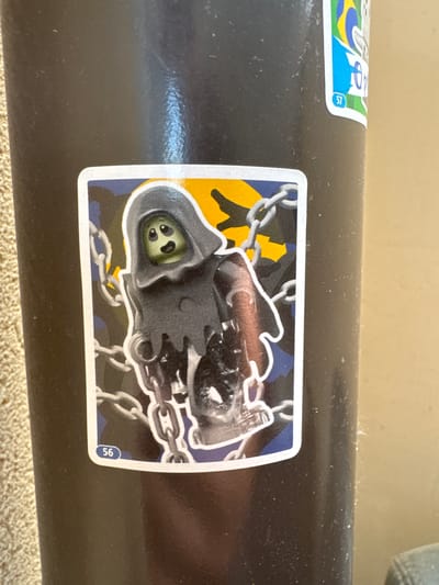 A sticker with a cartoon grim reaper is affixed to a dark, cylindrical object. The sticker is white-bordered and features a grim reaper wearing a hooded robe, with a yellow and black camouflage pattern in the background. The grim reaper's face is light green with two black eyes and a small black mouth. Chains are draped around the grim reaper. The number "56" is visible in a blue circle in the bottom right corner of the sticker. To the left of this sticker, a portion of another sticker is visible, which is white-bordered and features a blue, yellow, and green design with the number "57" in a blue circle. The cylindrical object appears to be a railing or pipe, possibly outdoors given the lighting and the textured surface beneath it. There are no specific indicators of time of day, weather, or strong emotions. The location context suggests this media file was captured in Suceava, Romania.