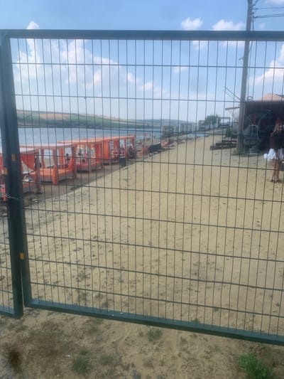 The image shows a sandy area with a body of water to the left, viewed through a wire fence. Several orange structures, possibly beach cabanas or tents, are set up on the sand near the water. People are visible within and around these structures, engaged in various activities. The sky is blue with some clouds, suggesting fair weather. The scene appears to be a beach or lakeside recreational area, likely during the daytime given the lighting. The sand is light brown, and there are some sparse patches of greenery. The overall impression is one of a relaxed outdoor setting.