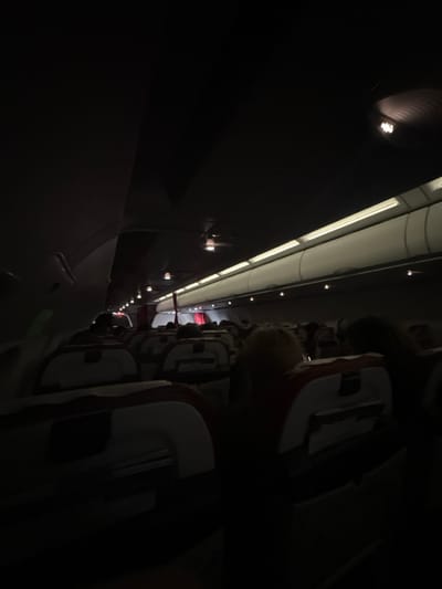 This image depicts a dimly lit commercial aircraft cabin interior, with the scene identified as being in Combs-la-Ville, France. The perspective is from a passenger seat, looking forward down the central aisle. Overhead bins line the cabin ceiling, which features continuous strip lighting and several individual, recessed light fixtures. The ambient lighting is low, suggesting either an overnight flight or a preparatory phase. Rows of airline seats, characterized by light-colored frames and dark or red upholstered sections, are visible extending into the distance. Seatbacks include visible tray tables and literature pockets. Multiple passengers are seated throughout the cabin, primarily discernible as dark silhouettes or indistinct forms due to the low light conditions. No specific actions, interactions, or individual facial features are clearly visible.