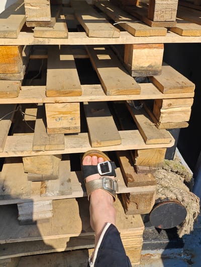 The image shows a person's foot wearing a sandal, placed on a stack of wooden pallets. The pallets are made of rough, weathered wood and are stacked in multiple layers. Some of the pallets have visible nails and grain patterns. The lighting suggests an outdoor setting with direct sunlight creating shadows. To the right of the pallets, a thick, rough rope and a dark, cylindrical object are visible. The background is dark and indistinct. The context provided that this image is located in Novonazimovo, Russia, suggests a possible outdoor storage area or a workshop in that location.