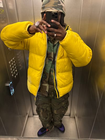 An individual is depicted taking a mirror selfie within an elevator in Savigny-sur-Orge, France. The subject wears a camouflage-patterned beanie, a bright yellow puffer jacket partially unzipped, a camouflage-patterned shirt, and matching camouflage cargo pants. Dark blue sneakers with black laces are on their feet. The subject's face is obscured by a gold-framed smartphone, held by the right hand. A large, oval-shaped ring with a pale purple stone is visible on the right index finger, and a silver-colored band ring is on the left ring finger. The elevator interior consists of highly reflective metallic walls and floor. A control panel with a grid of multiple square buttons and a rectangular, illuminated digital display is mounted on the left wall.