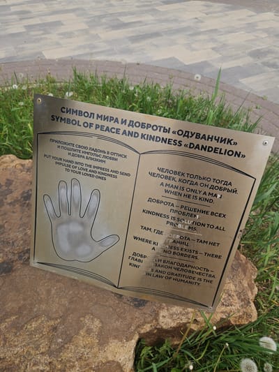 The image shows a metal plaque titled "Symbol of Peace and Kindness 'Dandelion'" written in both Russian and English. The plaque includes text about kindness, such as "A man is only a man when he is kind" and "Kindness is solution to all problems." It also depicts a handprint. The plaque is mounted near the ground, and partially covered by grass and resting on what appears to be a textured rock or pathway. The scene is in an outdoor setting with dandelions in the background. Based on additional information, the location of the image is in Речицы, Russia.
