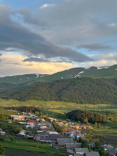 This is an aerial view of the village of Fioletovo, Armenia. The village is nestled in a valley at the base of green, rolling mountains, some of which still have patches of snow on their peaks. The houses are clustered together, with a mix of traditional stone and wooden buildings, many with tin roofs. Some houses have small gardens or agricultural plots around them. The village is surrounded by lush green fields and dense forests. In the distance, a line of power poles runs across the landscape. The sky is partly cloudy, with dramatic clouds tinged with the warm colors of sunset or sunrise, suggesting the photo was taken during the golden hour. There are no people visible in the image.