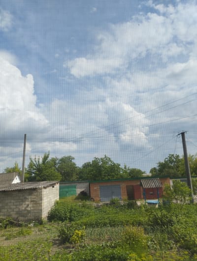 The image shows a view of several brick garages and a cinder block shed under a partly cloudy blue sky, with power lines running horizontally across the frame. In the foreground, there is dense green vegetation and what appears to be a garden. To the left, a white house with a grey roof is partially visible behind the shed. A concrete utility pole stands to the left, and another to the right, with several wires attached. The garages are varied in color, with some featuring metal doors and others brick or colored facades. A blue-covered well is situated in front of one of the garages. The scene is set in a rural or semi-rural area.