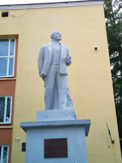 A monument of Vladimir Lenin stands in front of a building with yellow walls. The monument is made of light gray material and depicts Lenin in a suit, with his right hand holding a book. The building has several windows, some of which are partially visible on the left side of the image. There is also a small flag mounted on the wall to the right of the monument. Two security cameras are visible on the wall, one near the top of the building and another lower down on the left side. The monument has a plaque on its base with Russian text. The background features the branches of a tree. The overall scene suggests an outdoor setting on a bright day. This monument is located in the city of Faliukovo, Russia.