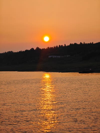 This media file captures a serene sunset over a body of water, likely a river or lake, near Потапово, Russia. The sun, a bright orb of orange, is low on the horizon, casting a warm, golden glow across the sky and the rippling water. A reflection of the sun shimmers on the surface of the water, creating a dazzling path of light.
In the background, a dark, silhouetted treeline forms a natural border against the vibrant sky. Perched on the hill behind the trees, several small, light-colored structures are visible, possibly buildings or shelters, adding a subtle human element to the otherwise natural landscape. The overall mood is peaceful and tranquil, evoking the quiet beauty of a day coming to a close. The time of day is clearly evening, with the warm colors of the sunset indicating fair weather. There is no visible text in the image.