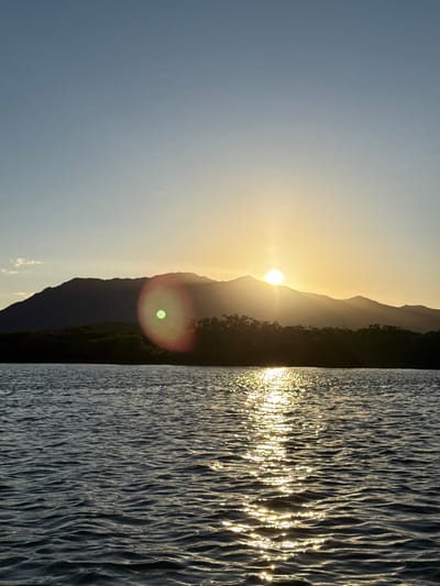 The image presents a sunset scene over a body of water in Dumbéa, France. The sun, a bright yellow disc, is positioned low on the horizon, partially obscured by the dark silhouette of undulating mountains in the background, primarily on the right side of the frame. A distinct green circular lens flare with a reddish halo is visible to the left of the sun. A dark band of dense vegetation, consistent with mangroves, forms the shoreline between the mountains and the water. The lower half of the image features calm, dark blue water with numerous ripples, reflecting the golden sunlight in a prominent, shimmering path extending from the horizon towards the foreground. The sky transitions from a clear light blue at the top to a bright yellow-orange near the sun. No individuals, man-made objects, or specific actions are discernible.