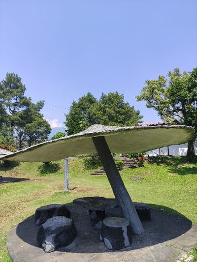 The image displays an outdoor scene featuring a concrete shelter with a mushroom-shaped canopy supported by a single, thick, angled concrete pillar. Beneath the canopy, on a circular concrete base, there is an irregularly shaped concrete table and at least five dark, cylindrical concrete stools designed to resemble natural stone or tree stumps.
The ground surrounding the seating area is covered in green grass. In the background, a variety of trees with green foliage are visible, including tall evergreen trees on the left and broader deciduous trees on the right. A faint outline of a distant hill or mountain is present on the horizon under a clear, bright blue sky. A slender, light gray metal pole stands vertically in the mid-ground to the left of the main shelter support. To the right in the background, a partial view of a white building or wall with a dark roofline is visible through the trees, along with a set of ascending concrete steps on a grassy slope.
No individuals are present in the scene. There are no discernable actions or interactions depicted. The image contains no explicit visual information such as landmarks, signage, or unique architectural styles to confirm its location in the city of Staufenberg, Germany.