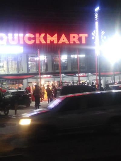 A nighttime shot captures the brightly lit facade of a "QUICKMART" store. Red illuminated lettering spells the store's name above the building. The store has multiple entry and exit points. Several people are gathered in front of the entrance. Cars are parked nearby, including one with headlights on in the foreground. The scene appears active and bustling, despite the darkness.