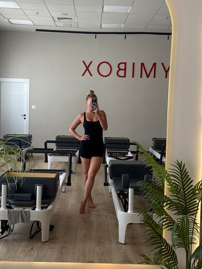 A woman stands in a Pilates studio, taking a mirror selfie. She is wearing a black athletic outfit and has her hair in a bun. The studio is modern and clean, with several Pilates reformers arranged in rows. A large logo is visible on the wall behind her, which reads "XOYMIB" in red lettering. The floor is made of light-colored wood, and there are plants in the foreground. The image is taken in Ariel, Palestinian Territories.