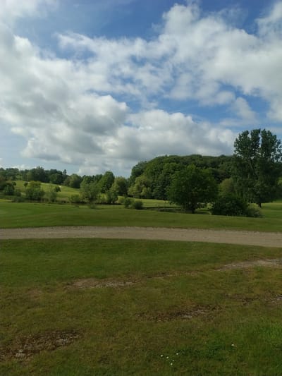 The image depicts a landscape in Lenningen, Luxembourg. A partially cloudy, blue sky dominates the left side of the frame. The right side is primarily green grass, separated by a narrow, gravel path. A line of trees runs roughly parallel to the path, further separating the sky and grass. No people are visible.