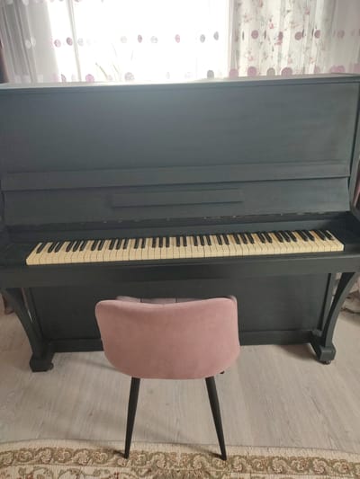A dark gray upright piano is positioned in front of a window with sheer curtains featuring a pattern of pink polka dots and small floral elements. A light pink velvet upholstered chair with dark slender legs is placed directly in front of the piano. The piano keys are visible, and some are slightly yellowed. The floor is light-colored laminate, and a rug with an intricate traditional pattern lies in the foreground. The scene appears to be an indoor setting, possibly a living room or music room, in Ivanovca, Russia. There are no people present in the image.
