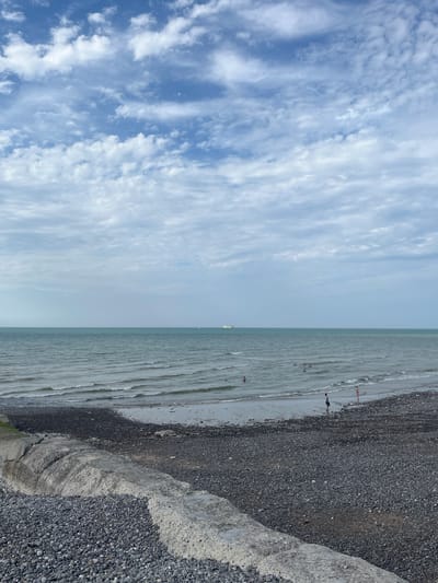 This media file captures a wide shot of a beach scene, split almost perfectly between a vast, cloudy sky and the ocean meeting the shore. The sky dominates the left side, filled with streaks of white and grey clouds against a pale blue background. These clouds appear wispy and thin, suggesting a bright but possibly overcast day.

The right side of the image presents the ocean, the beach, and a rocky embankment. The sea is a muted grey-blue, with small waves gently lapping at the shore. Scattered across the water and the beach are a few individuals. Some are wading or standing in the shallow water, while others are walking along the wet sand. The beach itself is a mixture of sand and dark pebbles, with a notable concrete or stone structure curving along the right edge, acting as a barrier or part of a promenade. This structure is textured with a rough, uneven surface. The far right of the image is covered in a dense layer of dark, small pebbles.

A small white boat is visible in the distance on the water, and a few distant figures suggest the beach is open to visitors. The overall atmosphere is serene and appears to be a casual day at the seaside. The lighting suggests it is daytime, likely late morning or afternoon, with diffused sunlight filtering through the clouds. The location context of Hautot-sur-Mer, France, places this scene on the French coast. There is no visible text in the image.