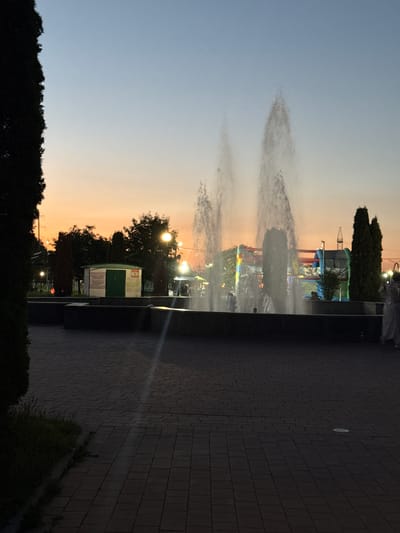 The image depicts a dusk scene in Endirey, Russia, likely in a public park or recreational area. In the foreground, a paved walkway of reddish-brown brick pavers is visible, leading towards a large, dark, rectangular fountain structure. Several plumes of water are actively spraying upwards from the fountain, illuminated by ambient light.
Behind the fountain, the environment transitions to a grassy area with scattered trees and bushes, silhouetted against the twilight sky. A small, white, shed-like building with a green door and a green roof stands prominently among the trees. Several streetlights are visible, casting pools of yellow light on the ground and illuminating pathways and gathering areas.
Further in the background, on the right side of the image, an inflatable attraction, possibly a bounce house or playground structure, is present, featuring bright colors like blue, yellow, and red. A crane is also visible in the far distance. A few individuals are present in the scene, appearing as indistinct figures due to the low light conditions, with one person on the far right dressed in light-colored clothing.
The sky above displays a gradient of colors, transitioning from a pale blue at the top to warm oranges and yellows near the horizon, indicating the time of sunset. On the far left of the image, a tall, dark, coniferous tree stands out, its dense foliage almost completely obscuring the sky. The overall atmosphere is peaceful and suggests a public space winding down for the evening.