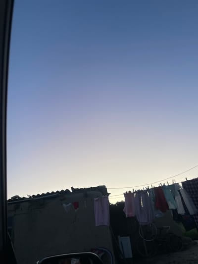 The image depicts a clear blue sky with a gradient transition to a lighter shade near the horizon, suggesting it is either sunrise or sunset. In the foreground, a dark, curved edge suggests the view is from within a vehicle. Along the right side of the frame, a building with a corrugated roof is visible, silhouetted against the bright sky. Clothes are hung on a clothesline strung across the building, with several garments of various colors, including pink, red, and white, visible. A bicycle is also present near the building. The scene conveys a sense of everyday life in what appears to be a rural or semi-urban setting. The overall impression is one of a tranquil and perhaps humble environment. The provided location context indicates the scene is Kara-Oi, Kyrgyzstan.