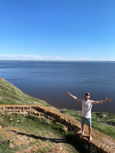 A low-angle shot depicts a light-skinned person standing barefoot on a weathered brick structure adjacent to water. The person wears a light-colored t-shirt, dark shorts, and a hat, and holds their arms outstretched. The water body fills the middle portion of the image, with the clear blue sky occupying the left side. The brick structure is topped with grass and vegetation. The photograph was taken in Шуран, Russia.