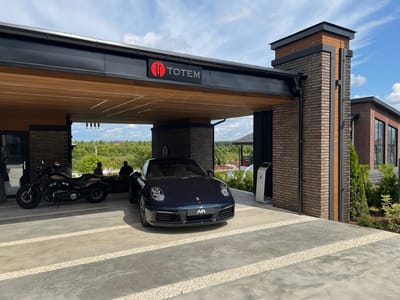 In Yakunino, Russia, a dark blue Porsche 911 is parked under a modern entryway structure. The entryway features a black metal roof with wood paneling on the underside and a "TOTEM" logo. The structure is supported by brick columns. To the left of the Porsche, a black motorcycle is parked. Two indistinct figures stand near the motorcycle, and another figure is visible in the background. The ground is a concrete driveway with white gravel accents. The scene is illuminated by natural daylight under a partly cloudy sky.