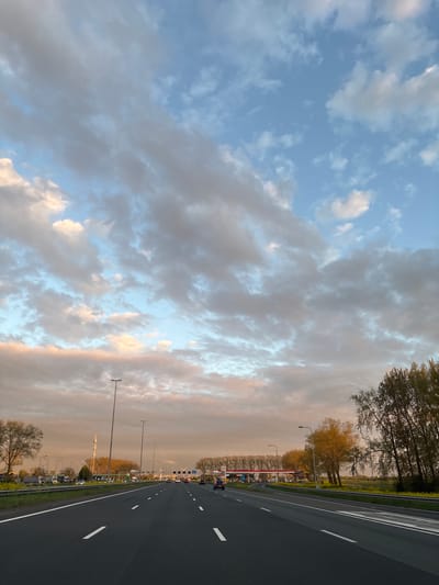 The image presents a road extending into the distance, possibly a highway, with trees on the right and a cloudy sky. The road is asphalt, and white lines mark the lanes. Street lights line the right side of the road. Further in the distance on the right, there appear to be buildings, including gas station. The sky is filled with clouds. Given the prompt includes "Beesd, Netherlands," it's possible this scene is located on a road in that Dutch town.