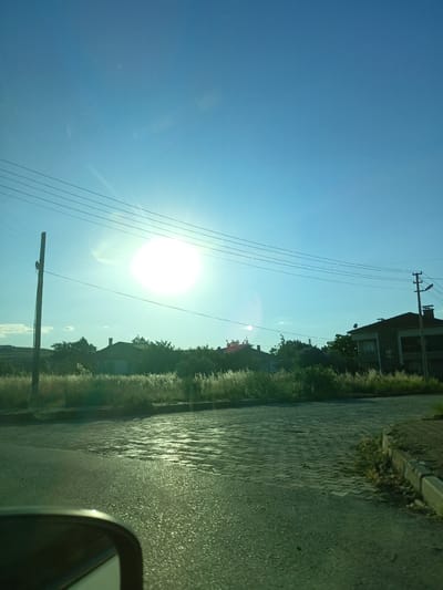 The image captures a bright, sunny day in Bahşılı Belediyesi, Turkey. The sky is a clear, vivid blue, with the sun appearing as an intensely bright, almost blinding, white orb in the upper left quadrant of the frame. Several power lines are strung across the sky, supported by a utility pole on the left side of the image. In the middle ground, there is a row of houses with dark roofs, surrounded by lush green trees and tall, dry grass that catches the sunlight. The foreground shows a paved road with a reflective surface, suggesting it may be wet or recently cleaned. A portion of a car's side mirror is visible in the bottom left corner of the frame, indicating the photo was taken from inside a vehicle.