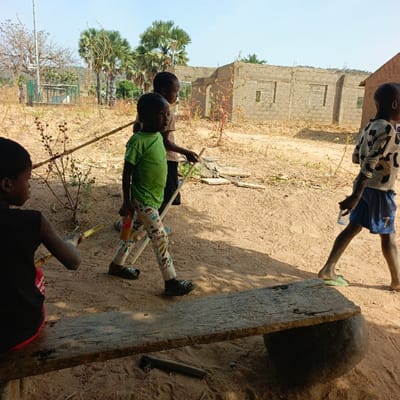 The image depicts an outdoor scene in Belnin, Nigeria, under a clear blue sky. The ground is unpaved, consisting of light brown, sandy earth with sparse, dry vegetation.
In the foreground, a dark, weathered wooden plank serves as a bench, supported by a large, dark, spherical object. A dark, cylindrical object rests on the ground beneath the bench. To the left, a child in a dark sleeveless top with a red stripe is seated on this bench, facing right and holding a yellowish object.
In the mid-ground, three other children are present. A child in a bright green t-shirt, patterned long pants, and dark sandals walks from left to right, holding a long stick in their right hand and a small bottle in their left. Slightly behind and to their right, another child in a light-colored t-shirt also walks left to right, holding a stick. To the far right, a fourth child wearing a patterned long-sleeved shirt and blue shorts with a '7' on the leg walks right to left, carrying a clear plastic bottle.
The background features several unfinished structures constructed from grey concrete blocks, with visible door and window openings. To the left of these buildings, several tall palm trees with green fronds are prominent. A tall utility pole with equipment is visible in the distant left background. The scene is brightly lit by direct sunlight, casting shadows.