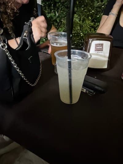 The image depicts a dimly lit outdoor setting, likely a cafe or bar, in Seregno, Italy. In the foreground, a clear plastic cup contains a light-colored beverage with ice and a black straw, situated on a dark tabletop. To the left, a black purse with a silver chain strap is visible, and a person's hand is partially obscuring the purse's zipper. To the right of the cup, a brown dispenser with a white logo featuring the letter 'M' is present. Behind the dispenser, another person's arm and hand are visible. In the background, lush green foliage suggests an outdoor seating area. Another partially visible plastic cup with a yellowish beverage sits behind the cup in the foreground. A black rectangular object, possibly a cigarette pack or phone, lies on the table near the clear cup.