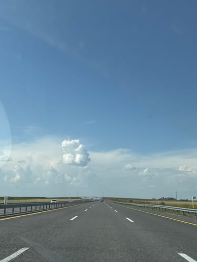 A straight, multi-lane highway stretches into the distance under a vast blue sky dotted with white clouds. The road is paved with asphalt and has clear white lane markings and a yellow line along the right edge. Guardrails line both sides of the highway, separating it from grassy fields on either side. In the distance, a white car is visible in the right lane. Further down the road, a highway sign indicates "142". The environment appears to be rural or undeveloped, with a flat landscape. There are no people clearly visible. The overall scene depicts a travel or journey along an open road on a clear day. The image is captured from a moving vehicle, as suggested by the perspective. The specific location in Алтынды ауылдық округі, Kazakhstan, cannot be definitively determined from the visual information alone, but the landscape suggests a potentially open and vast area common in such regions.
