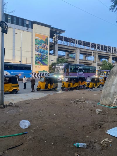 This image captures a street scene in St. Thomas Mount-Pallavaram Cantonment Board, India, under an overcast sky, likely during dawn or dusk. The foreground is a muddy, uneven ground littered with debris, including a crumpled plastic bag, a green hose, and a plastic water bottle. A dirt path leads towards a paved sidewalk.

Beyond the sidewalk, a queue of yellow auto-rickshaws, typically used as taxis, is lined up. Two men are standing near the auto-rickshaws; one in a white shirt and dark pants, and another in a brown shirt and pants. A blue Ashok Leyland bus is parked further down the street, displaying the company logo.

In the background, a multi-level elevated structure, possibly part of a transportation network like a metro or flyover, dominates the scene. A large building with a mural depicting rural life and a stylized sun is visible on the left. A prominent purple bus with "CAPTAIN COACH" written on its side is also present in the traffic lane. The overall impression is of a busy urban environment with elements of transportation infrastructure, public transport vehicles, and everyday street life.