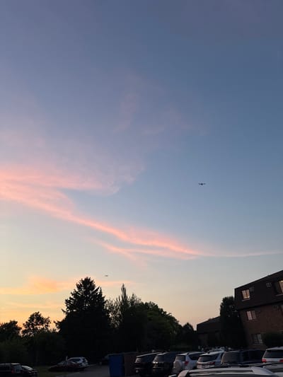 The image displays an outdoor scene at either sunrise or sunset in the city of Glendale Heights, United States of America. The sky exhibits a gradient of colors, transitioning from a muted blue in the upper portion to purples, light blues, and distinct bands of pink and orange clouds closer to the horizon. Two aircraft are visible as dark silhouettes against the lighter sky: one prominent aircraft in the upper-right quadrant, and a smaller aircraft in the lower-left portion, situated above the tree line.
The ground level is dominated by a dense treeline composed of both coniferous and deciduous trees, forming a dark silhouette across the mid-ground. In the foreground and extending into the mid-ground, a paved parking lot is occupied by numerous parked vehicles, primarily SUVs and sedans, appearing dark due to the low light conditions. On the right side of the frame, a multi-story brick apartment building is visible, featuring a dark, sloped roof with dormer windows and multiple rectangular windows on its brick facade. A blue dumpster is positioned in the parking lot adjacent to the building. No individuals or direct interactions are visible within the frame.