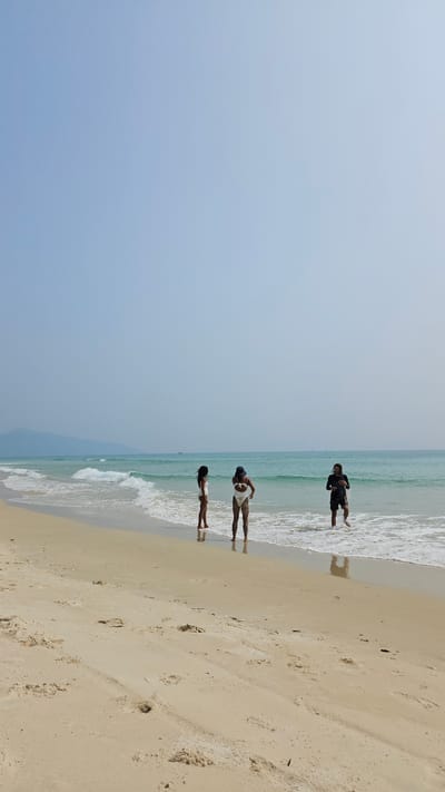 A vibrant, sunny beach scene captured at Bac Cam Ranh, Vietnam, shows a clear blue sky meeting a tranquil ocean and a sandy shore. The image, when viewed in its intended landscape orientation, features a wide expanse of light blue sky at the top, transitioning to the clear, turquoise waters of the sea, and a wide strip of light-colored sand along the bottom.

Three individuals are visible along the water's edge. One person, dressed in dark clothing, is positioned furthest from the viewer, standing in the shallow ocean water with their back partially turned. Closer to the camera, another person wearing light-colored attire and a hat stands in the water, appearing to look down. The third individual, closest to the foreground, is standing right where the gentle waves meet the sand, also dressed in light clothing. All three cast clear reflections on the wet, shimmering sand.

The ocean exhibits small, white-crested waves gently lapping at the shore. The beach consists of fine, golden sand, with various footprints and small debris scattered across it. The bright, even lighting across the scene indicates a clear day, likely midday or early afternoon, with no clouds visible in the sky. The overall impression is one of a peaceful and leisurely day by the sea. There is no visible text in the image.
