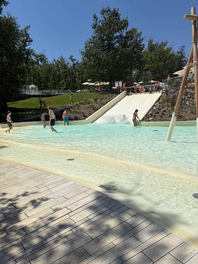 The image depicts a scene at a water park in Cavriglia, Italy. In the foreground, a wooden deck area leads to a shallow, clear pool of water with a sandy-colored bottom. The water is illuminated by sunlight, creating rippling reflections. Several people are visible in the pool, wading and interacting. To the right of the pool, a large white water slide descends from an elevated platform, with a person in a bikini at the bottom of the slide, appearing to exit the water. Further up the slide, a group of people are gathered, some preparing to slide down. The background features a grassy slope with trees and more foliage. In the distance, there are lounge chairs with umbrellas, suggesting a recreational area. The sky is clear and blue, indicating a sunny day. The water park appears to be an outdoor facility, integrated into a natural, wooded environment.