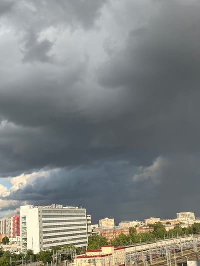 The image captures a dramatic sky with dark, heavy clouds dominating the upper portion, suggesting an impending storm or recent rainfall. Below the ominous sky, a cityscape unfolds. In the foreground, a prominent, multi-story white building with numerous windows stands out. To its left, several residential buildings, including one with red accents, are visible. Beyond these, a line of trees separates the buildings from what appears to be a railway station or industrial area with tracks and overhead structures in the lower part of the frame. The overall impression is an urban environment under a turbulent sky, characteristic of the weather that might be experienced in Veshki, Russia.