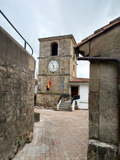The image presents an outdoor scene in Colunga, Spain, depicting a paved courtyard viewed from a narrow passage. In the left foreground, a stone-faced wall topped with a dark grey concrete section and a black metal handrail defines the passage boundary. On the right, a textured grey stucco building wall features a brown metal gutter system running vertically and horizontally, beneath a red-tiled roofline. The central and dominant structure is a multi-story, rough-hewn stone clock tower with a red-tiled roof and a cross finial. A circular clock face with Roman numerals is visible on the tower's facade, indicating approximately 12:40. Below the clock face, three flags are displayed horizontally: a white flag, a green/white/blue striped flag, and a red/yellow/red Spanish flag. Further to the left of these, another blue flag is visible. To the right of the clock tower, a white building with a red-tiled roof and a dark brown wooden door is accessed by a grey stone staircase lined with a black metal railing and potted plants. The courtyard ground is paved with rectangular light red and grey interlocking pavers. The sky is uniformly overcast. No people or animals are present.