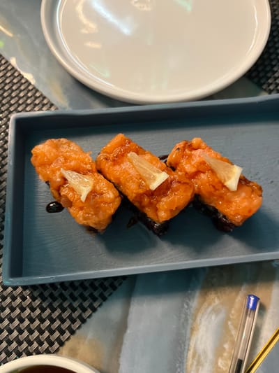This close-up, overhead shot captures a dining table setting, likely indoors. The surface features a dark, patterned placemat underneath a lighter, possibly marble-textured mat.

The central focus is a light blue or gray rectangular serving dish, holding three pieces of what appear to be baked or pan-fried sushi. Each sushi piece is topped with a generous portion of orange-pink salmon, which has a glossy, cooked appearance, and is garnished with a small, translucent wedge of lemon. Dark drizzles, possibly of a sauce like teriyaki or balsamic glaze, are visible beneath and around the sushi.

To the left, a portion of a clean white, oval-shaped plate is visible. In the upper right corner of the image, the end of a blue ballpoint pen can be seen. A small, partially visible white bowl containing a dark liquid, likely a dipping sauce, is situated in the lower right.

No people are present in the image. The arrangement suggests a meal is either about to be enjoyed or has just been served. The lighting appears to be indoor, possibly artificial, reflecting softly on the white plate. There is no visible text. While the context states the location is Basiglio, Italy, no specific visual cues within the image confirm this.