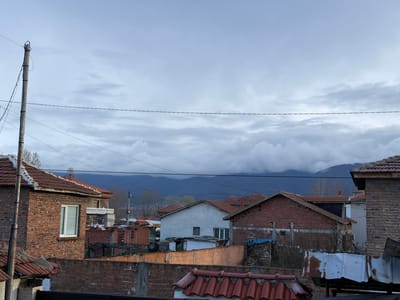 The scene depicts a cloudy sky above residential buildings in Liahovo, Bulgaria. The foreground consists of various rooftops covered with red terracotta tiles and brick walls of the surrounding houses. A utility pole with wires runs along the left side of the frame. In the middle ground, there is a mix of single and multi-story buildings, some brick and some stucco. Behind the houses, mountains are visible. The overall impression is of a quiet, overcast day.