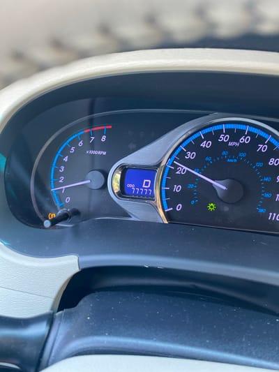 The image is a close-up shot of a car's dashboard, specifically the instrument cluster. A portion of the steering wheel is visible in the foreground. The dashboard features a speedometer with markings for MPH and Km/h, a tachometer, and a digital display showing "D" for drive and an odometer reading of "77777". Various warning lights and indicators are present, including a green light that appears to be for the headlights or running lights. The overall condition of the dashboard suggests it is in a vehicle. The lighting indicates it is daytime. There is no visible indication of the city of Prijeradi, Montenegro, or any people.