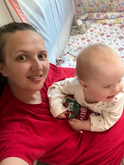 In an indoor setting, likely a child's crib or playpen, a woman and a baby are pictured. The woman is on the left side of the frame, her face close to the camera, and she is wearing a red t-shirt. Her expression is a mix of a smile and an open-mouthed gaze. Beside her, to the right, is a baby, also facing towards the right. The baby is wearing a light-colored onesie with a floral pattern and appears to be smiling and looking away from the camera. The background includes a white mesh netting on the left and a colorful bedding with hot air balloon and animal motifs on the right. A stuffed bunny toy is visible on the bedding. The overall scene suggests a moment of playful interaction between the woman and the baby. The location is identified as Maksimovo, Russia.