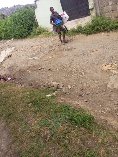 An outdoor scene in Kiamaina ward, Kenya, depicts a dusty, unpaved thoroughfare with scattered debris under natural daylight.

In the center, a young male, appearing to be in his late teens or early twenties, with short dark hair, walks from left to right. He wears a dark t-shirt, light-colored distressed or ripped trousers, and light-colored closed-toe shoes. He carries a white cardboard box with red markings in his right arm, and two nested plastic basins (one blue, one green) in his left hand. A light-colored bag or cloth bundle is slung over his left shoulder. His gaze is directed slightly downwards and to his right.

In the lower-left foreground, the lower leg and foot of another individual, wearing a pink open-toed sandal, are partially visible, obscured by a patch of grass.

The ground surface is predominantly dirt and gravel, uneven, with exposed rocks and various pieces of refuse, including torn paper/fabric and small plastic items. Patches of green and dry grass grow along the lower edges of the frame and a defined line separates the grass from the dirt path.

In the background, to the right of the central figure, stands a light-colored building with a large, dark-colored metal gate. Further right, a concrete wall is visible. To the left of the building, extensive green bushes and foliage cover a rising terrain. Beyond this, a distant hillside with sparse vegetation and faint structures is visible under an overcast sky.