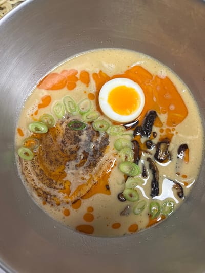 The image displays an overhead view of a prepared ramen dish contained within a silver-colored, likely stainless steel, bowl. The bowl holds a creamy, light tan broth, topped with numerous bright orange-red chili oil droplets and streaks. A soft-boiled egg, halved lengthwise to reveal a vivid orange-yellow, semi-liquid yolk, is positioned in the upper right quadrant. Multiple slices of fresh green scallions are scattered across the broth's surface. In the central-left, a textured, dark brown mass, consistent with seasoned ground meat or a dark paste, is visible. Several dark, cooked mushroom pieces are partially submerged in the broth's right half. No individuals are depicted. The image contains no discernible visual elements to confirm the specified location of Pozuelo de Alarcón, Spain.