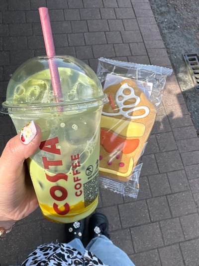 A hand holds a clear plastic cup containing a green beverage with ice and bubbles, topped with a domed lid and a pink straw. The cup has red lettering that reads "COSTA COFFEE." Next to the cup, also held by the hand, is a gingerbread cookie in a clear plastic wrapper. The cookie is decorated to resemble a house with white icing, yellow icing for a window frame, and orange icing with pink dots. The background is a paved surface made of dark grey rectangular bricks. The scene is outdoors, with sunlight casting shadows. The person is wearing blue jeans and black shoes with decorative floral embellishments. A black and white leopard print fabric is visible at the bottom of the image.